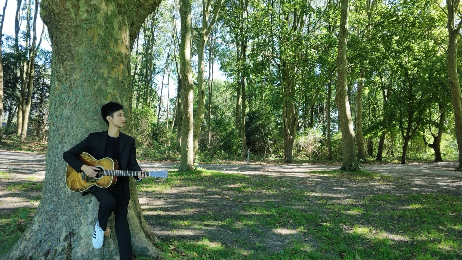 Jiasheng Hu holding an acoustic guitar beside a tree in a sunlit park