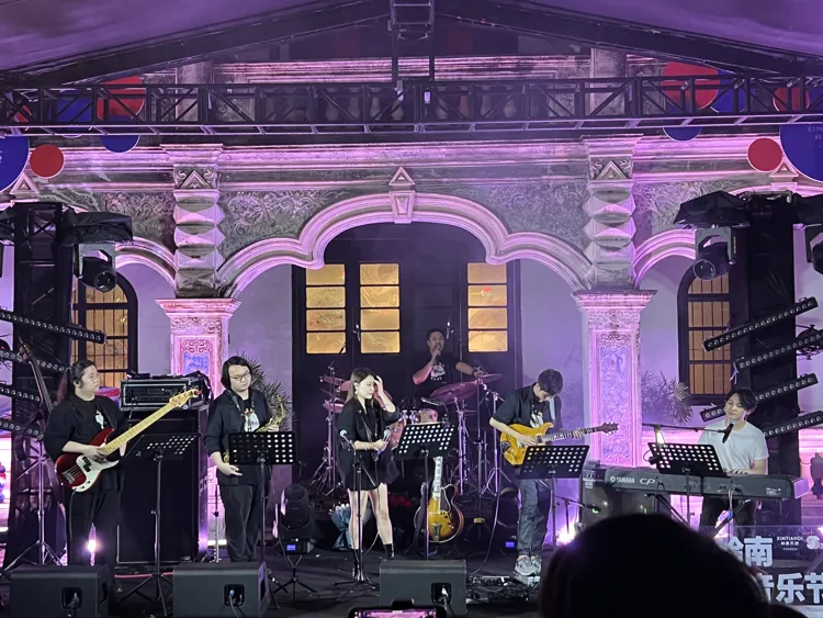 Full band performing on an outdoor stage with a purple-lit facade
